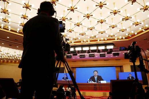 A silhouette of a reporter behind a TV camera recording a broadcast of a Chinese state official on a large screen.