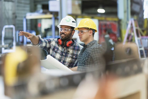 Two men in hard hats and protective glasses on a factory floor.