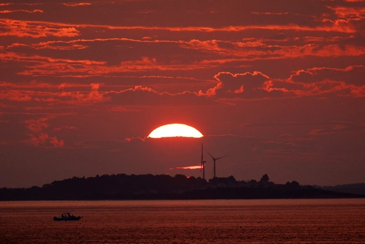 The sun sets behind a cloud and two wind turbines in front of a body of water.