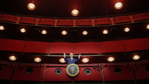 Donald Trump, wearing a blue suit, stands in a red theater box above the photographer with his hands raised.